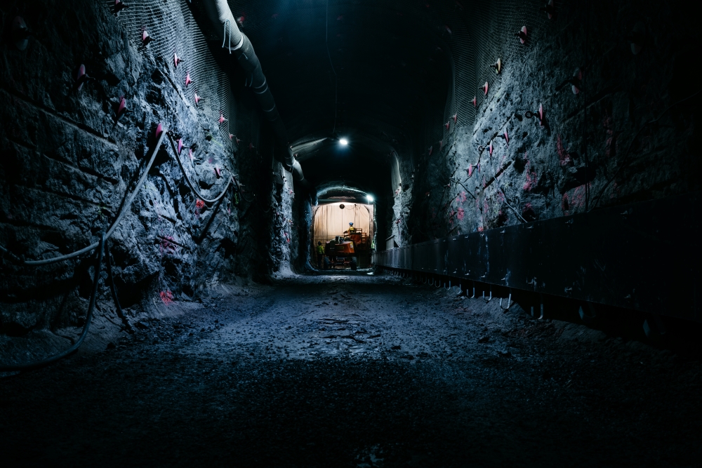 A dramatic shot looking down a dark underground rock tunnel secured with wire mesh, rock bolts, and survey markers. At the far illuminated end, workers in protective gear operate heavy mobile construc